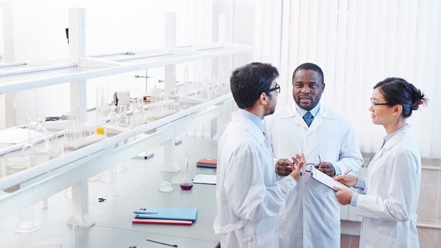 Three scientists in lab coats discussing their research in a lab with lab glassware on the shelf behind them.