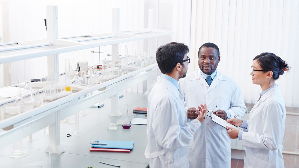 Three scientists in lab coats discussing their research in a lab with lab glassware on the shelf behind them.