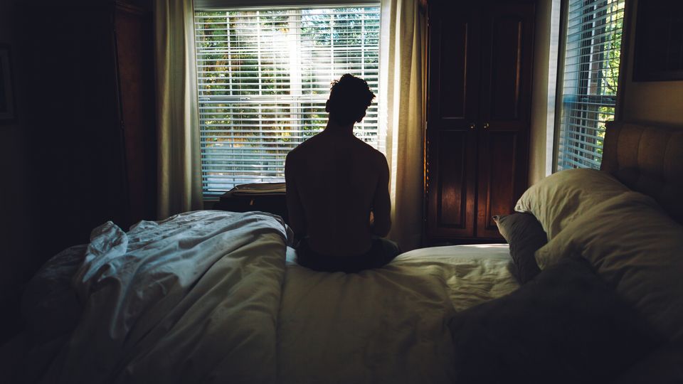 A man sitting on the edge of a bed, looking out of a window suggesting feelings of sadness.