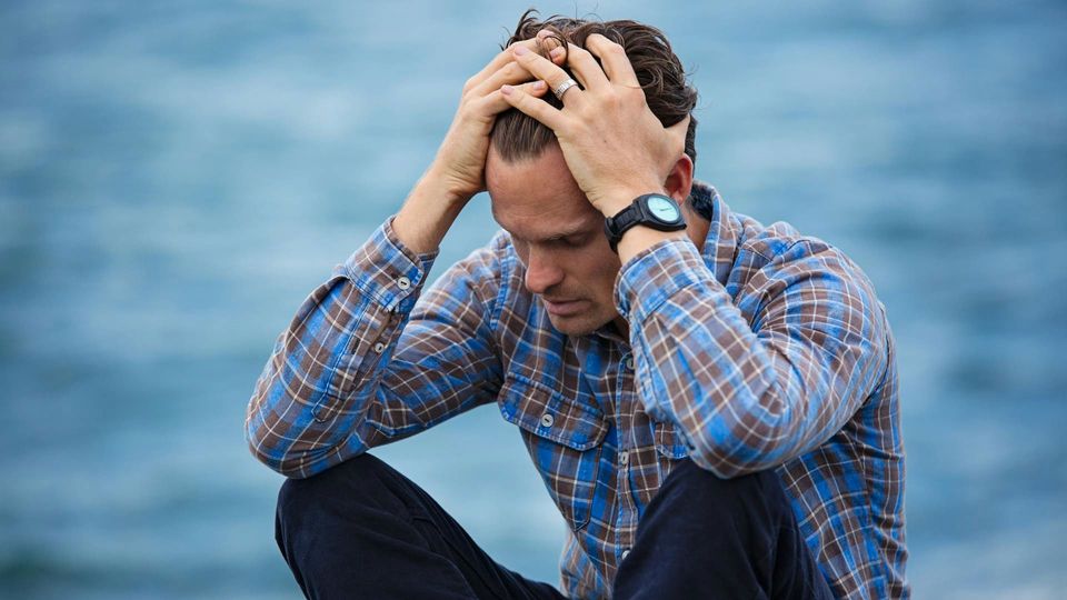 A man in a blue and brown plaid shirt runs his hands through his hair, looking stressed.