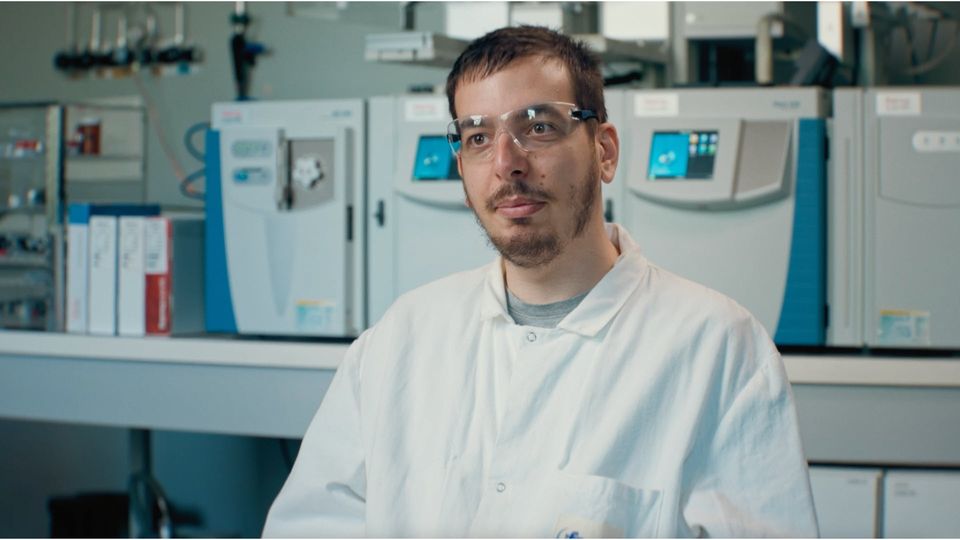 Headshot of a scientist with lab equipment behind him on a work desk