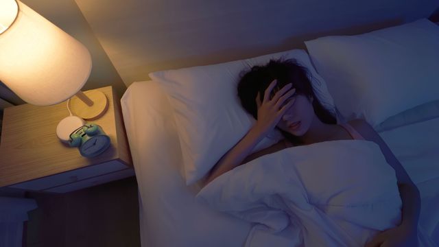 A woman in bed with a lamp on holding her head, suffering from poor sleep. 