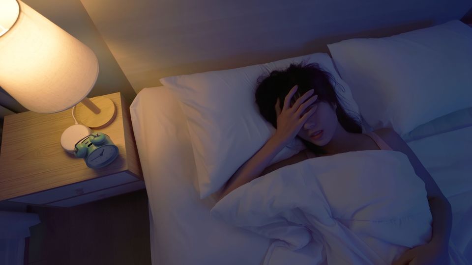 A woman in bed with a lamp on holding her head, suffering from poor sleep.