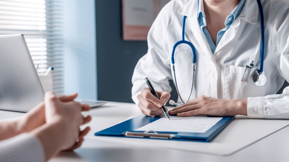 A doctor in a lab coat, wearing a stethoscope diagnosis a patient whilst writing notes on a notepad.