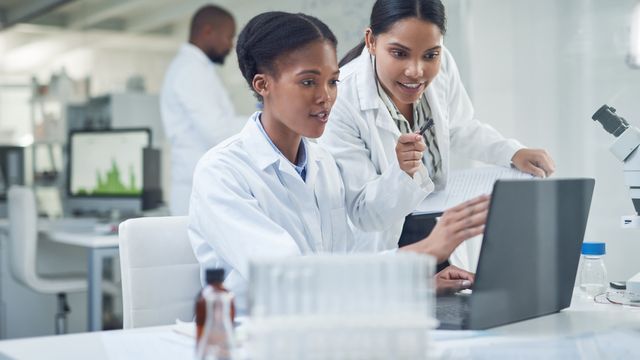 Two scientists in lab coats analyzing data on a laptop in a modern laboratory setting. 