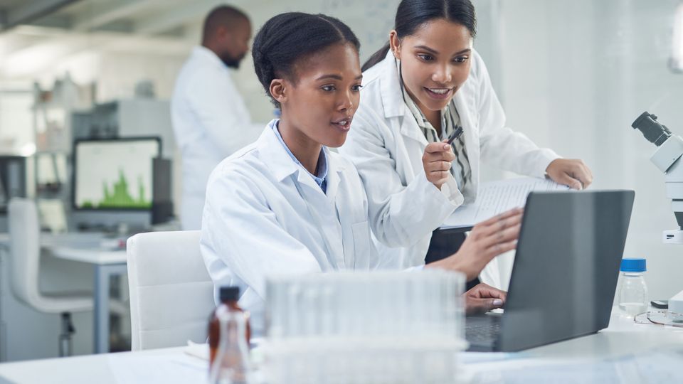 Two scientists in lab coats analyzing data on a laptop in a modern laboratory setting.