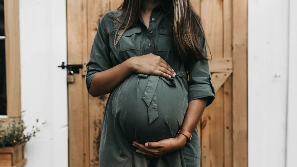A pregnant woman stands in front of a door, holding her bump in both hands.