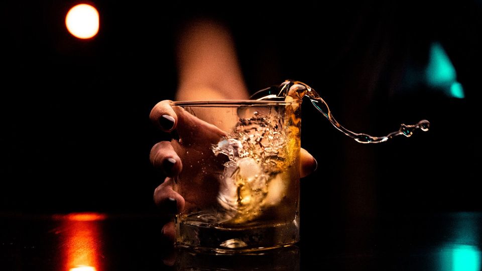 A person grabs a glass of alcohol, liquid spilling over the edge.
