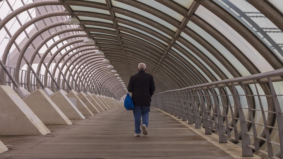 An older man walking away from the camera, down a bridge.