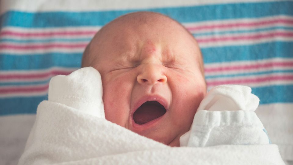 Newborn baby wrapped in a white blanket, yawning while lying on a striped hospital sheet.