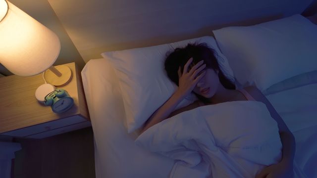 Woman lying in bed at night with her hand on her forehead, struggling to sleep, with a bedside lamp and alarm clock visible, representing insomnia. 