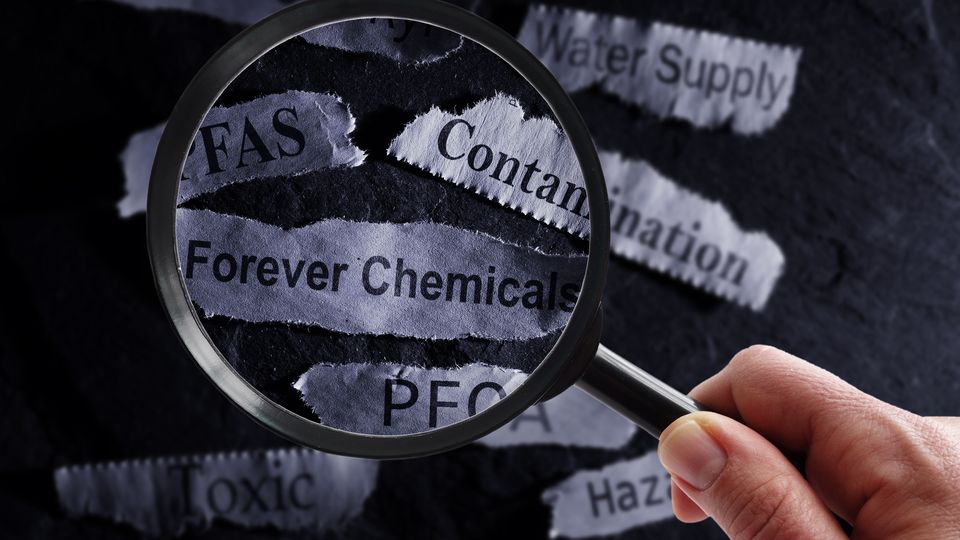 A person holding a magnifying glass over scraps of paper that read "PFAS", "Forever Chemicals", "Contamination"