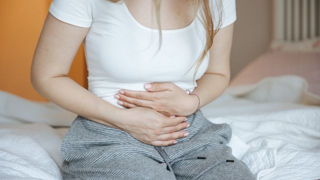Woman sitting on a bed holding her lower abdomen, showing discomfort from menstrual pain and cramps. 