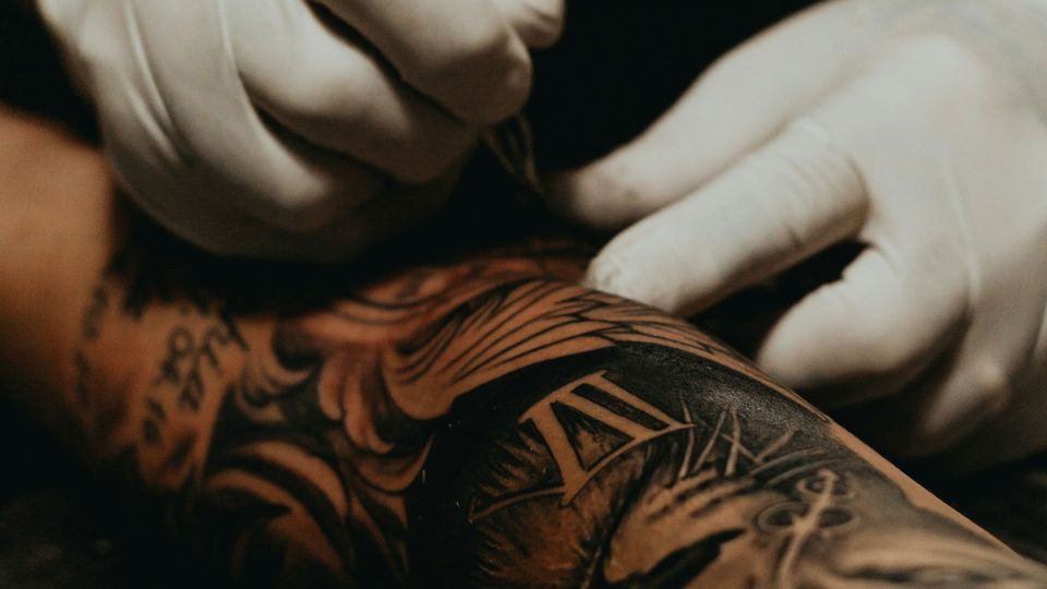 White-gloved hands tattoo black ink on a person's arm.