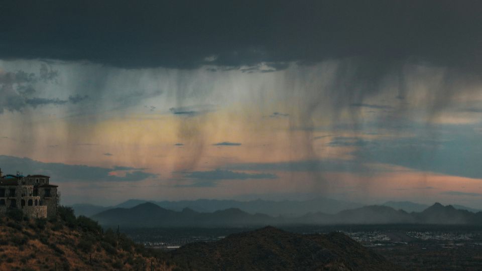 Spindles come down from a heavy rain cloud to pour on the distant land below. Mountains visible in background.