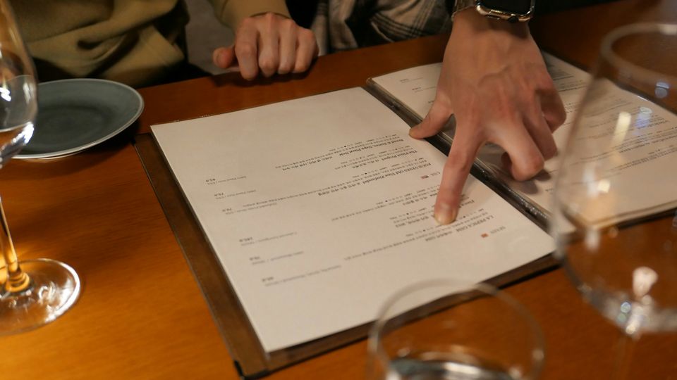 A hand pointing out options on a menu, which is resting on a dining table.