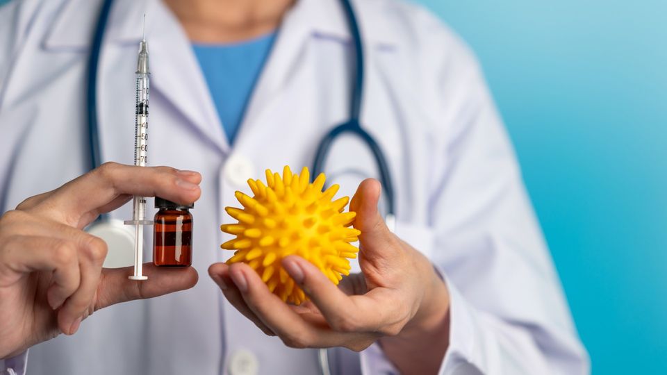 A doctor holding a vial of medicine and a needle in one hand, and a model of a virus in the other.