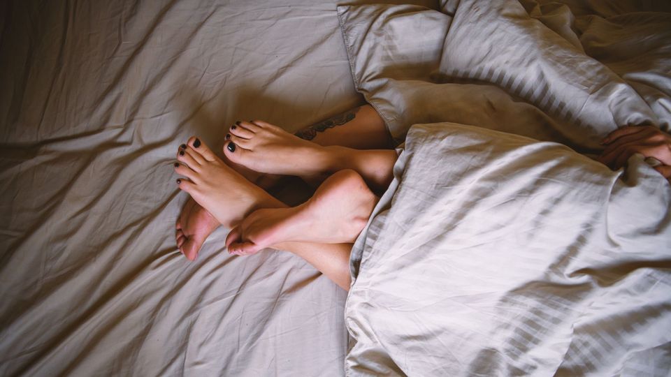 Couple’s intertwined feet under white sheets, symbolizing intimacy and virginity themes.