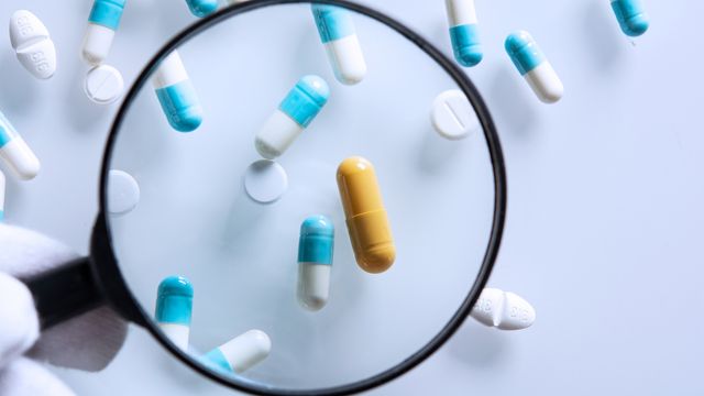 Magnifying glass highlighting a yellow capsule among various blue and white pills during drug screening. 