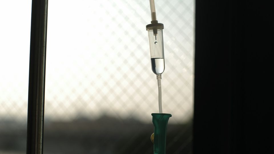 Clear IV drip chamber with fluid suspended in front of a window with a safety mesh pattern.