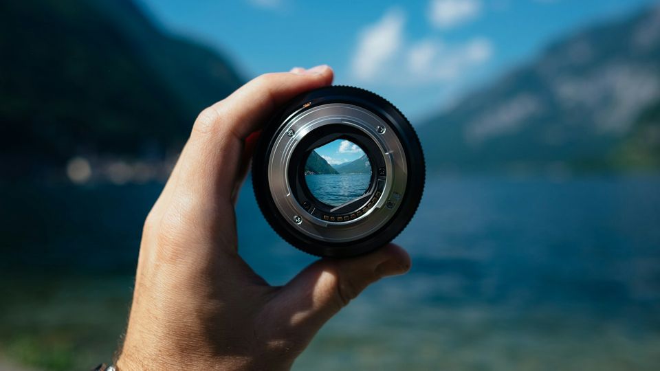Hand holding a camera lens with a sharp lake view inside, symbolizing vision focusing.