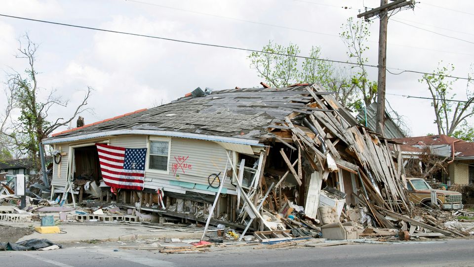 A wrecked wooden building, its structure collapsing into the street. American flag still clinging to its side. Hurricane devastation. 