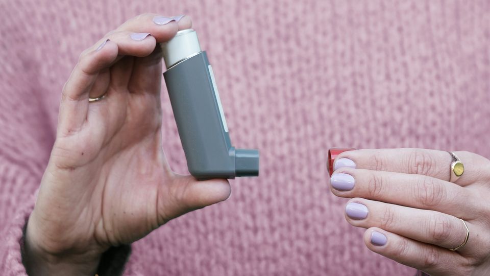 A woman in a pink knit jumper holding a gray asthma inhaler. 