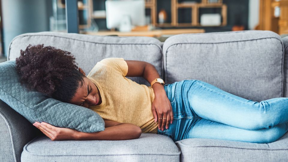 Woman lying on a couch holding her abdomen, showing signs of chronic pain and discomfort.