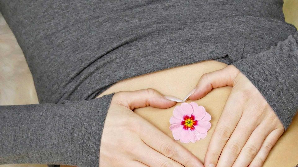 White woman holding her abdomen, hands positioned into an open heart shape with a flower nestled inside.