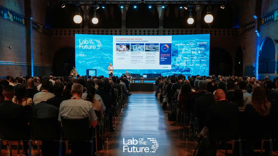 A speaker presenting at the Lab of the Future Europe Congress, with a packed room of audience members. Focal point is the presentation deck with a darkened room of audience heads.