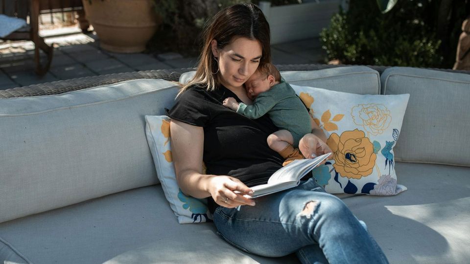 A mom reads a book while her baby sleeps on her.