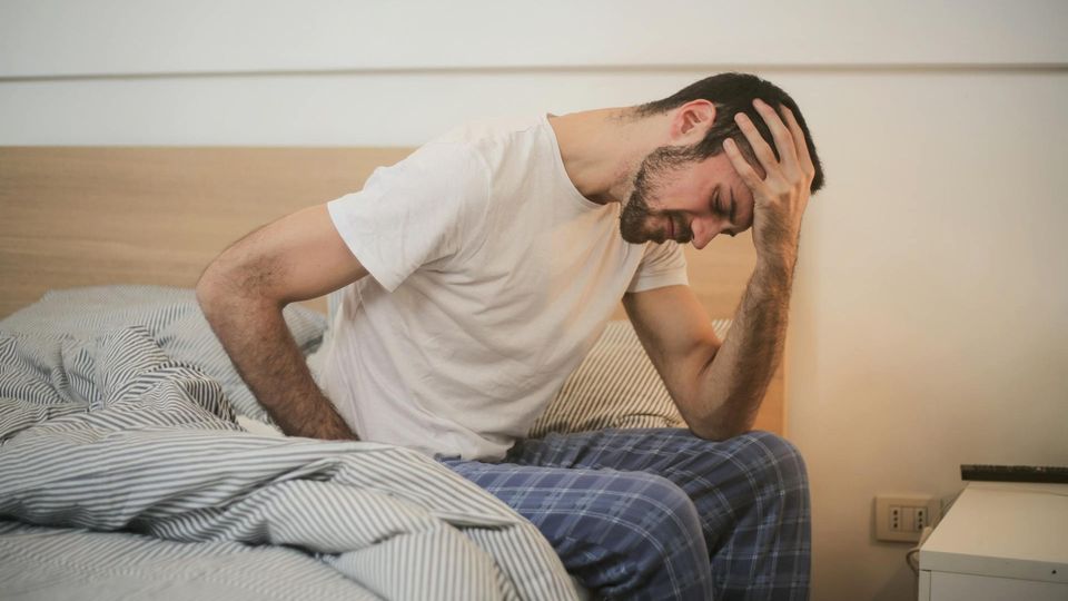 A man in pyjamas sits on a bed, doubled over in pain from a stomach ulcer. 