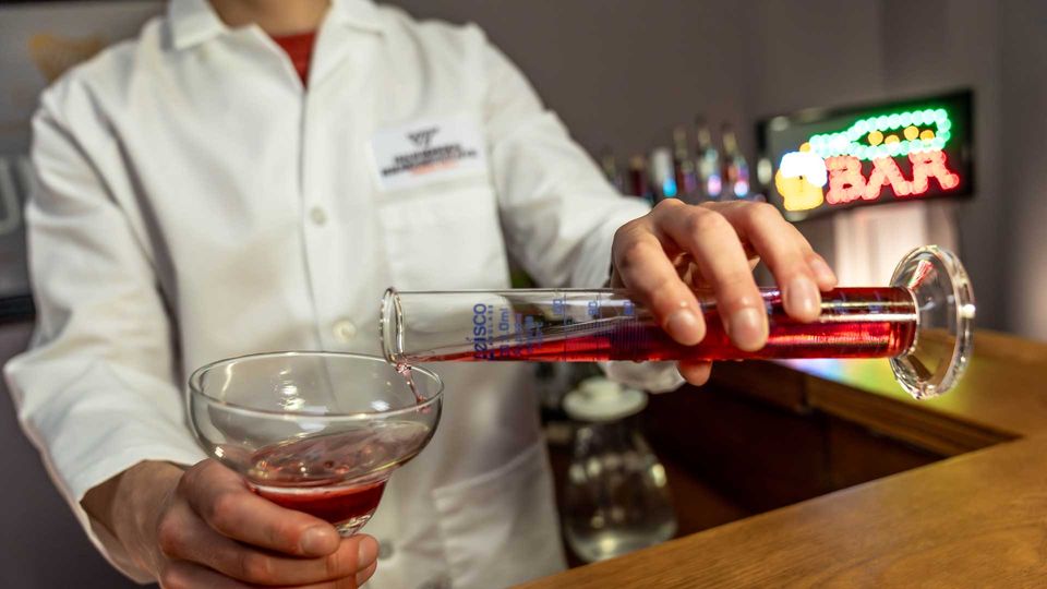 A person in a lab coat pouring a red alcoholic beverage into a cocktail glass, behind a bar.