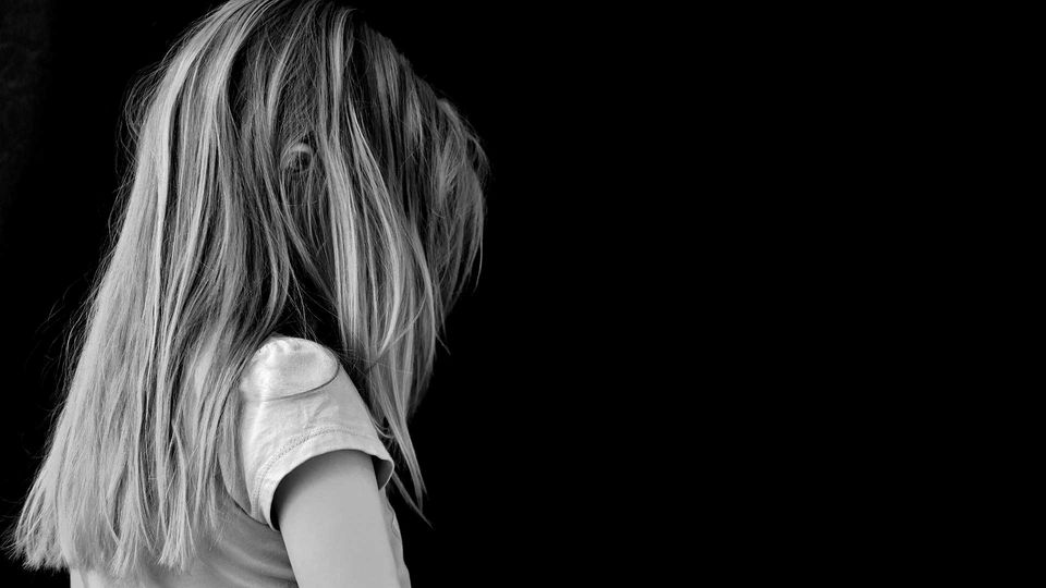 Photograph of a young girl with her back turned away from the camera, head down, against a black background. 