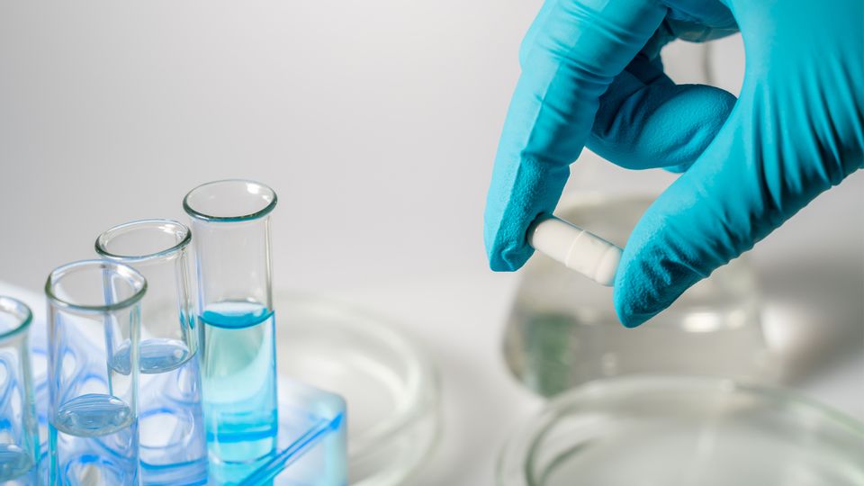 A gloved hand holding a pill next to test tubes containing blue liquid, representing proteomics in drug discovery.