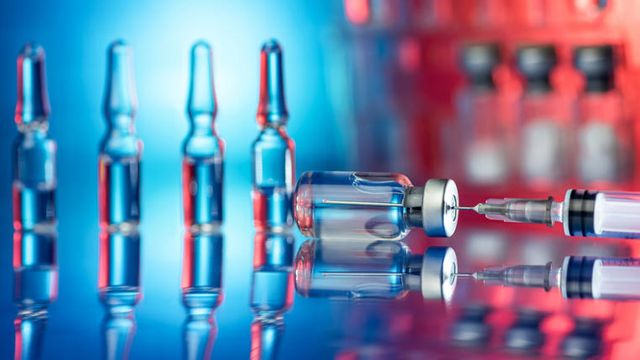 Close-up of vaccine vials and a syringe with reflections on a glossy surface under blue and red light 