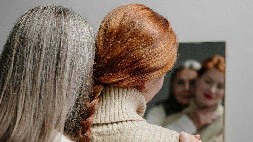 A woman with gray hair and a girl with ginger hair look in a mirror.