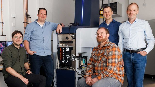 Five male researchers surrounding a modified modified fluorescence microscopy–t-MALDI-2 mass spectrometer, smiling. 
