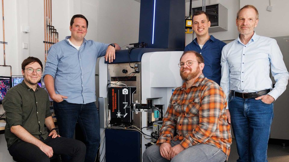 Five male researchers surrounding a modified modified fluorescence microscopy–t-MALDI-2 mass spectrometer, smiling.