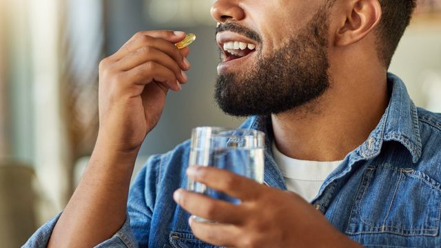 A man holding a glass of water in one hand, lifting a vitamin supplement pill to his open mouth. 