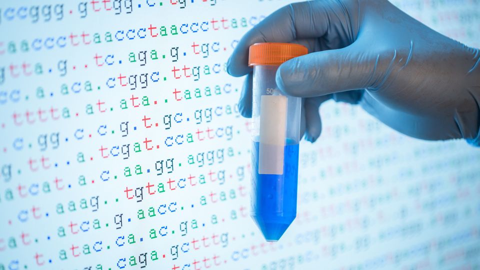 Gloved hand holding sample tube in front of DNA sequence data used in next-generation sequencing.