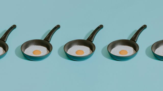 A plain blue background on which five small frying pans are sitting in a row, each containing a fried egg. 