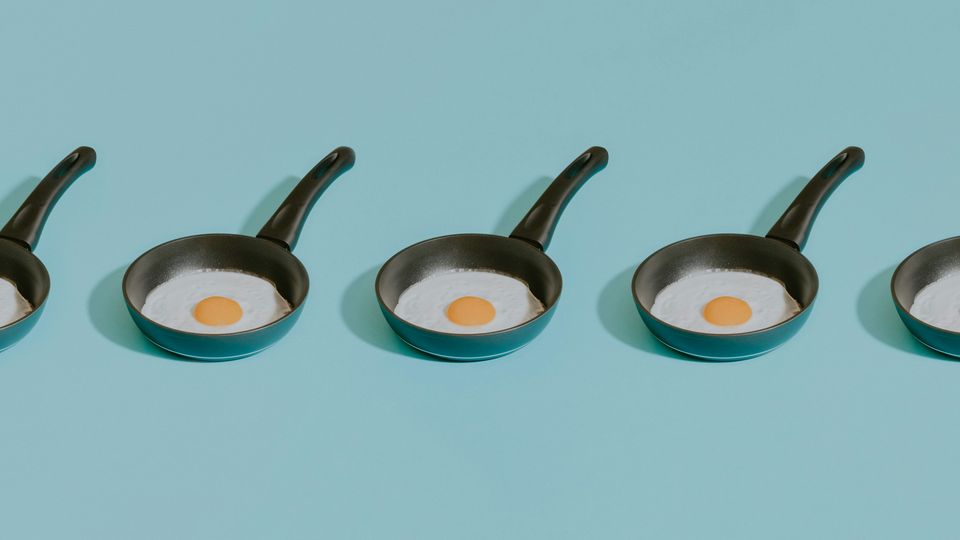 A plain blue background on which five small frying pans are sitting in a row, each containing a fried egg.