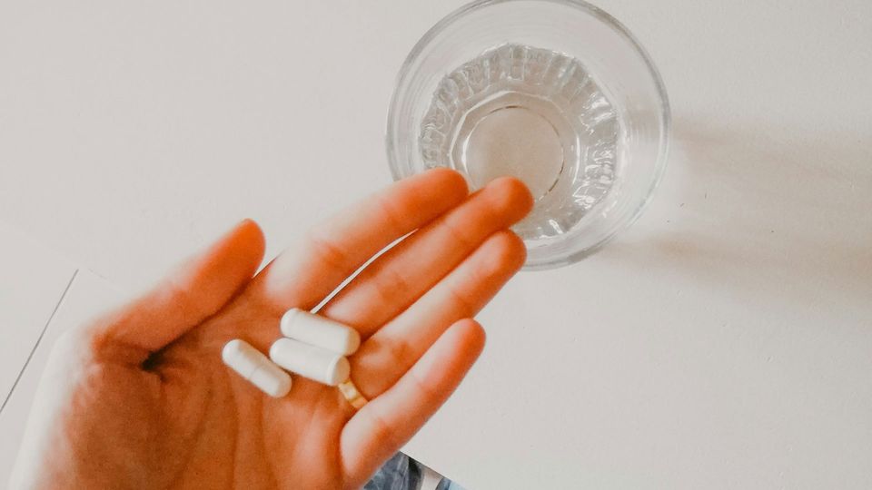 Three white capsules rest on a person's hand, with a glass of water on a table in the background.