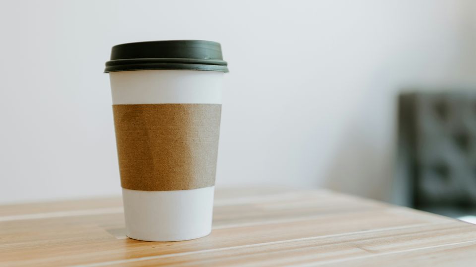 A paper coffee cup with a black plastic lid and brown cardboard sleeve, sitting on a wooden table.