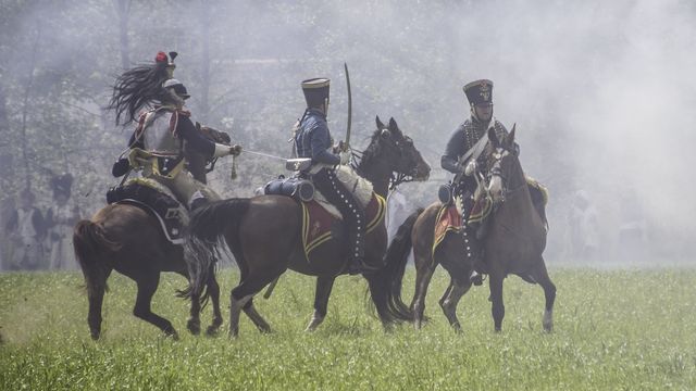Historical reenactors dressed as Napoleonic cavalry soldiers riding horses through battlefield smoke. 