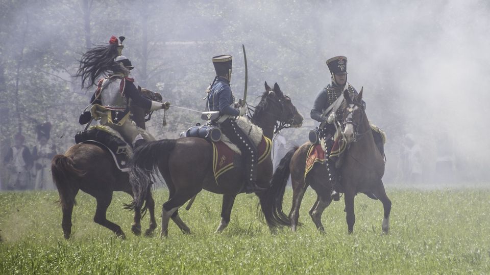 Historical reenactors dressed as Napoleonic cavalry soldiers riding horses through battlefield smoke.