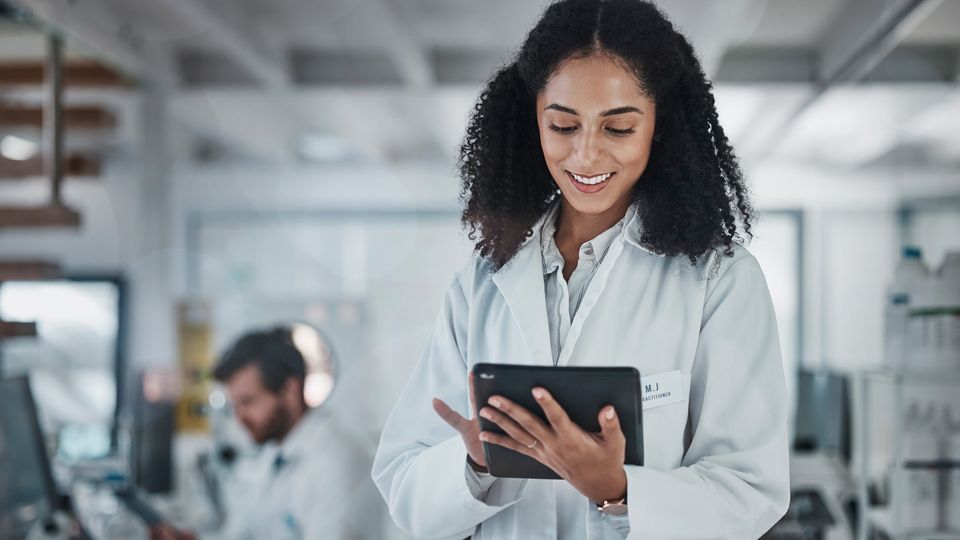 Female scientist in a lab coat smiling while using a digital tablet in a modern laboratory setting, with a colleague working in the background.