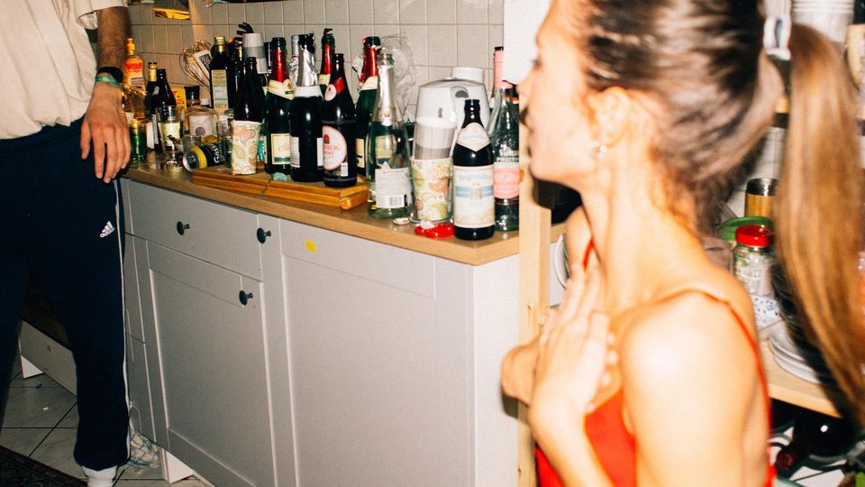 Two people at a house party in a kitchen with numerous bottles of alcoholic beverages on the counter, suggesting social drinking.