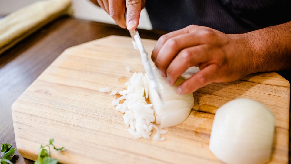 A man dicing a white onion with a chef's knife on a wooden chopping board.